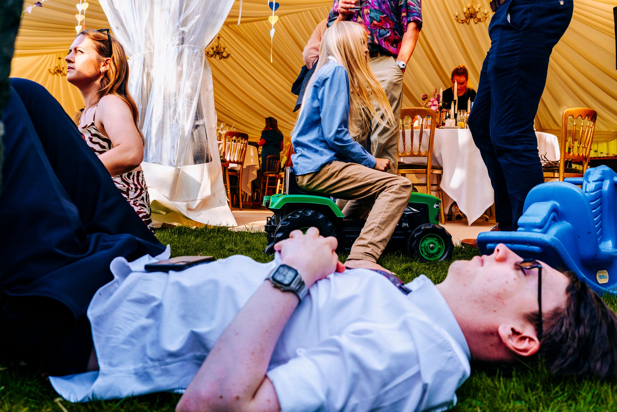 Wedding guest lying on grass near marquee while children play with toy tractor at outdoor reception.