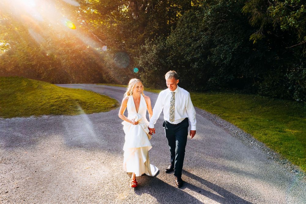 Bride in white dress and groom in tie walk hand in hand on gravel path in golden sunlight.