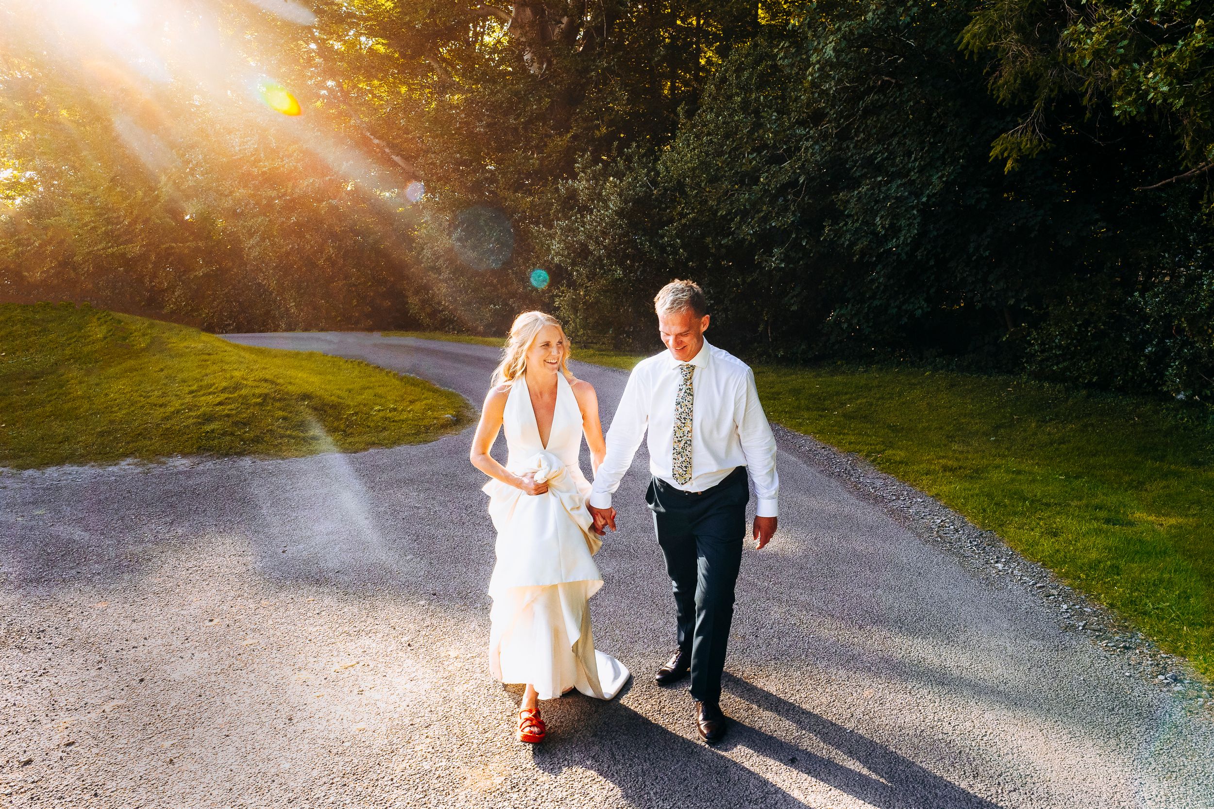 Bride in white dress and groom in tie walk hand in hand on gravel path in golden sunlight.