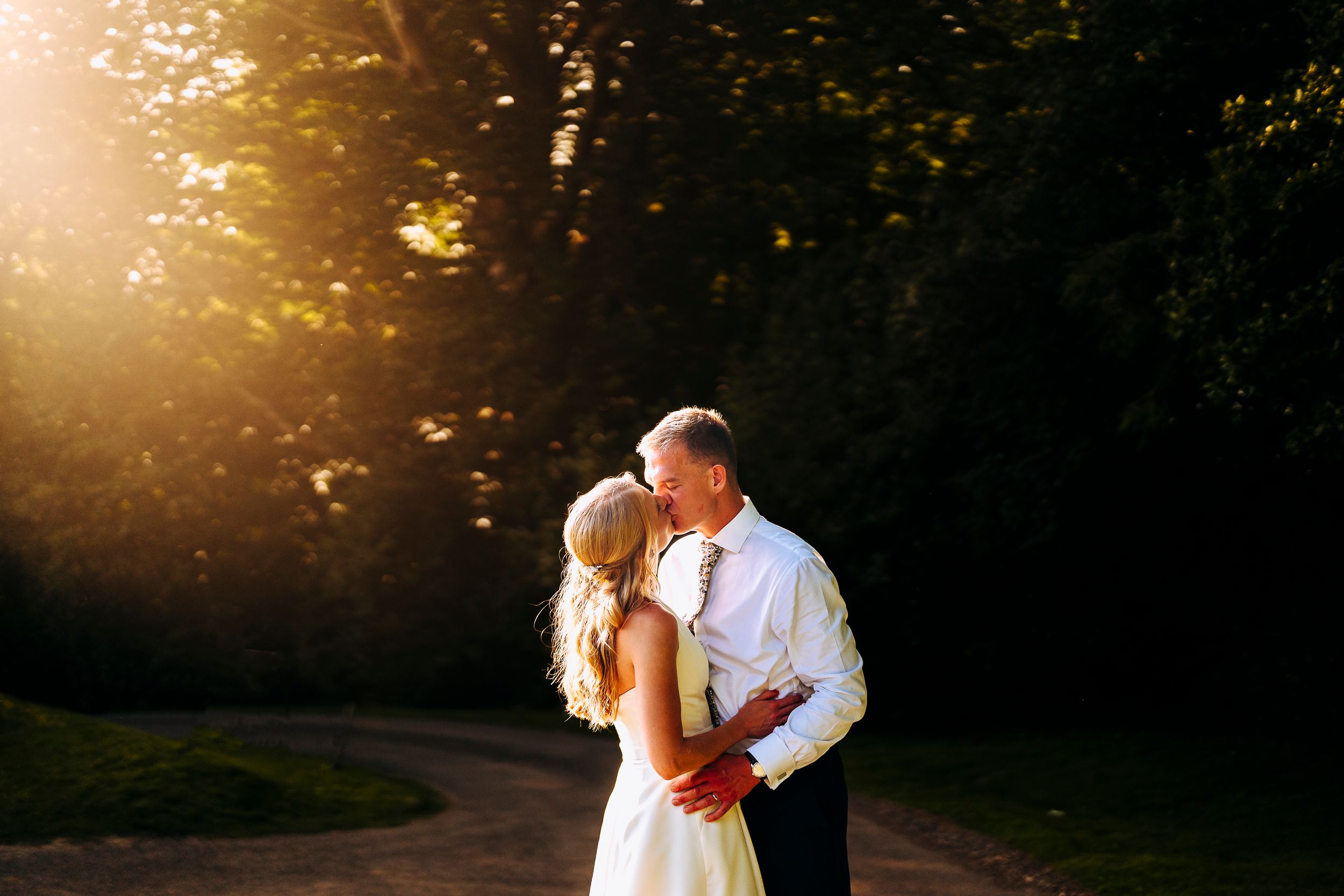 Bride and groom sharing romantic golden hour kiss surrounded by lush green trees at wedding.