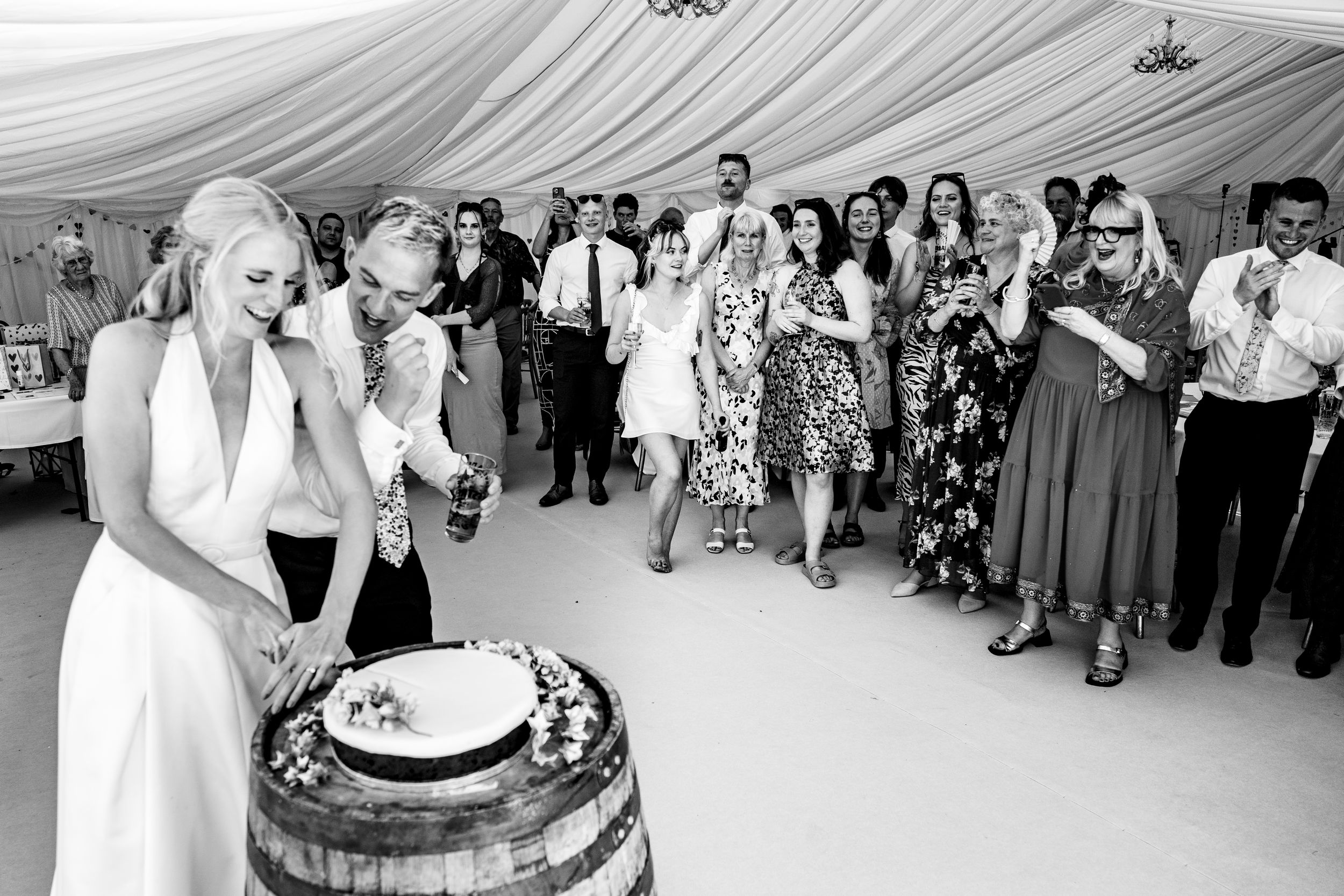 Bride cutting wedding cake on barrel in black and white photo with guests watching in marquee.