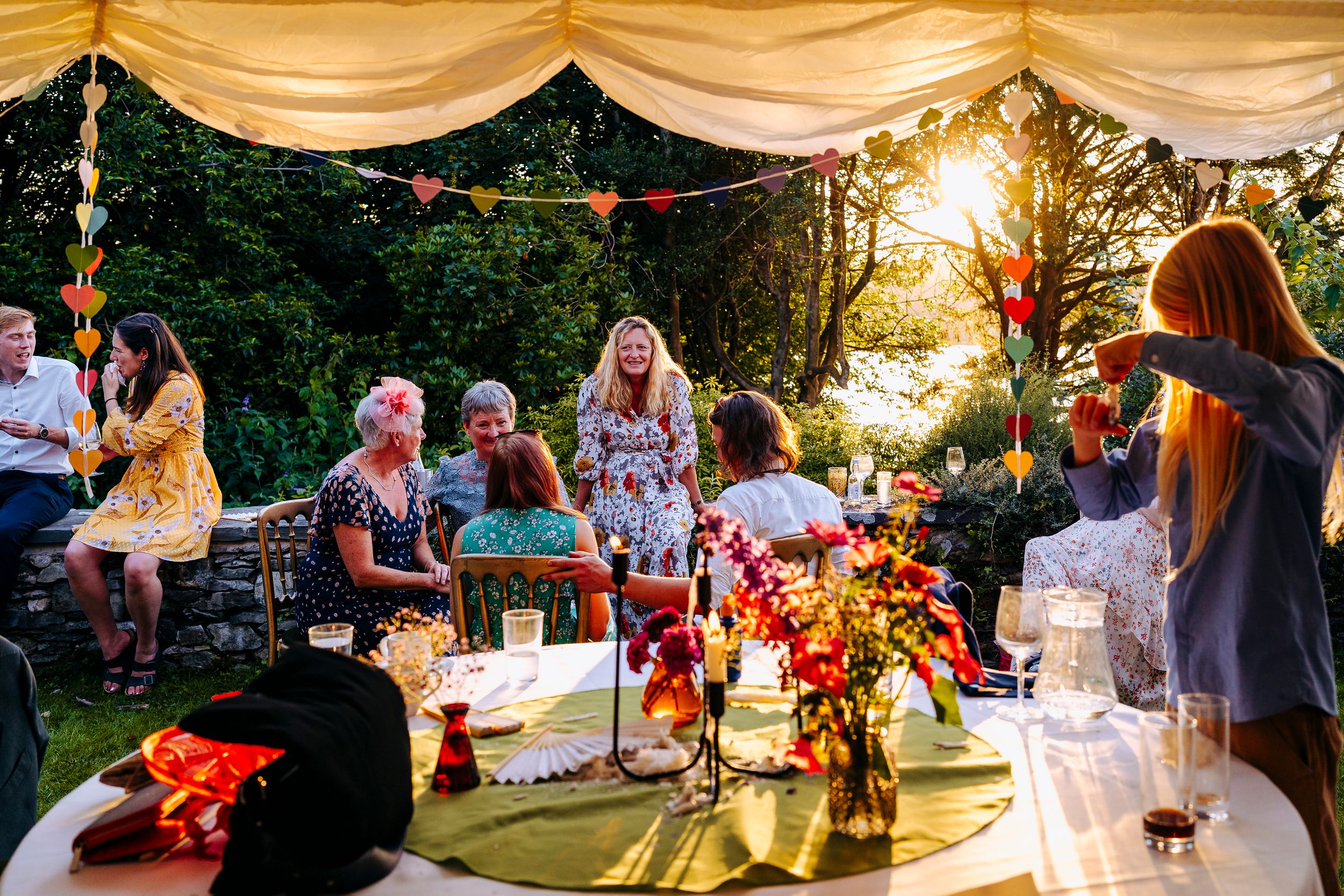 Wedding guests gathered around table in golden sunset light beneath decorated outdoor marquee.