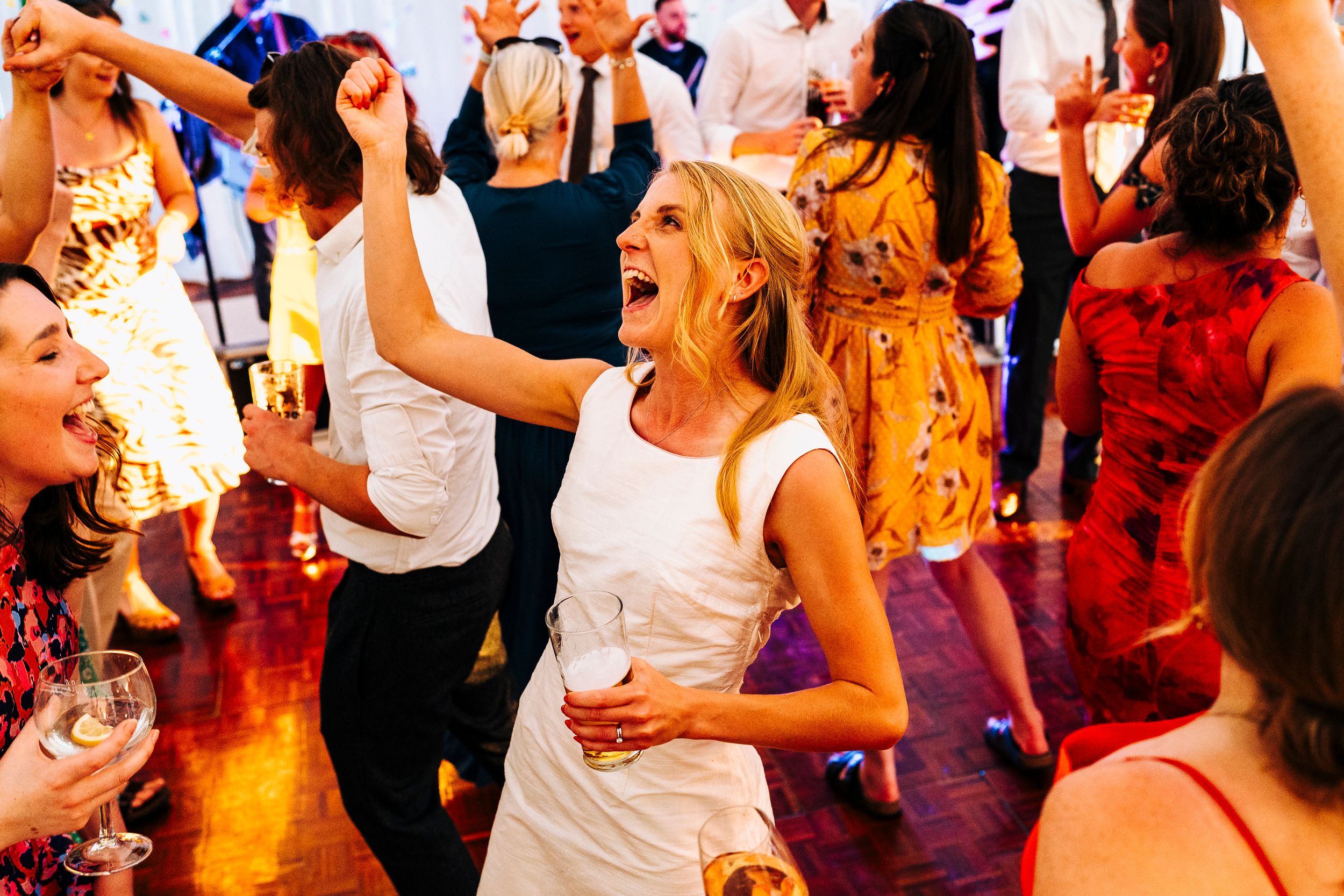 Radiant bride in white dress dancing joyfully with drink in hand at lively wedding reception.