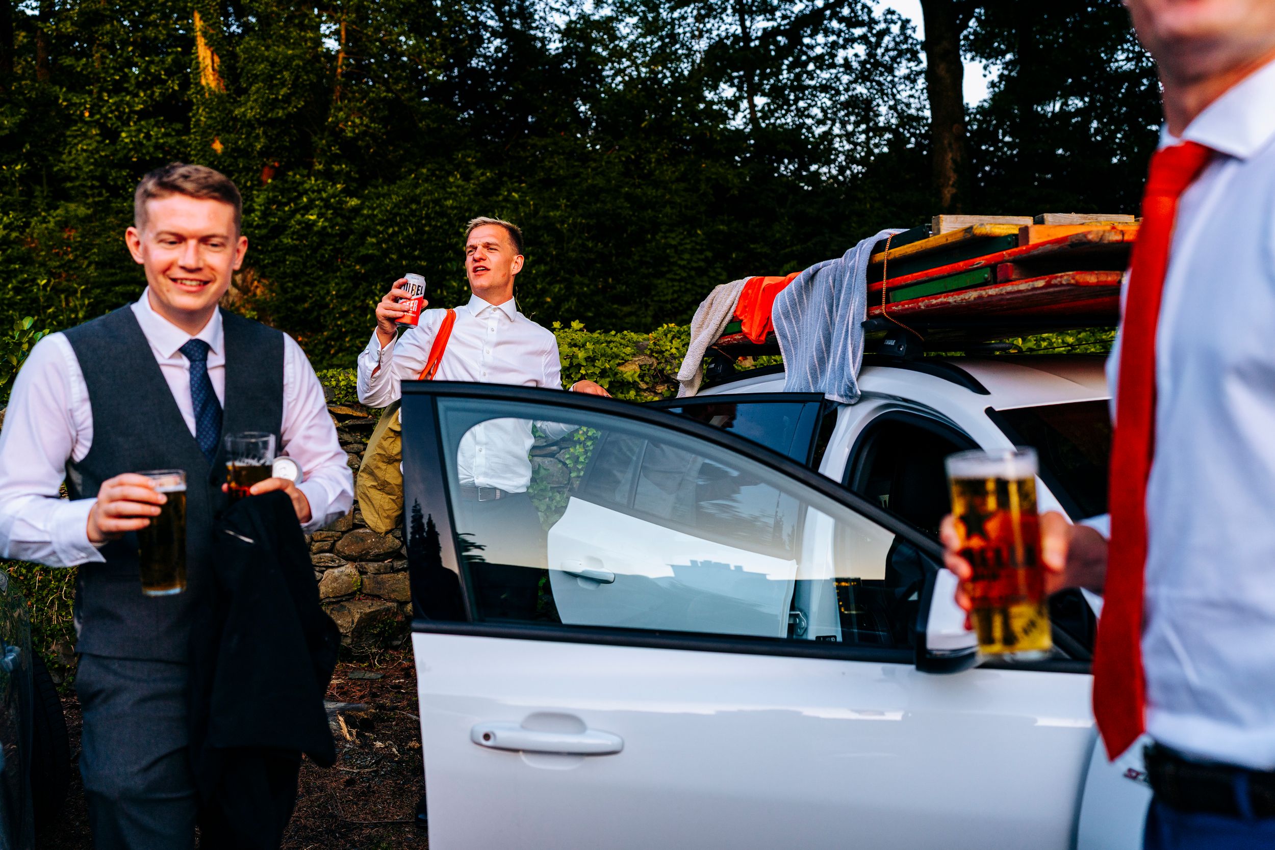 Smartly dressed wedding guests smiling and socializing beside white car at outdoor venue.