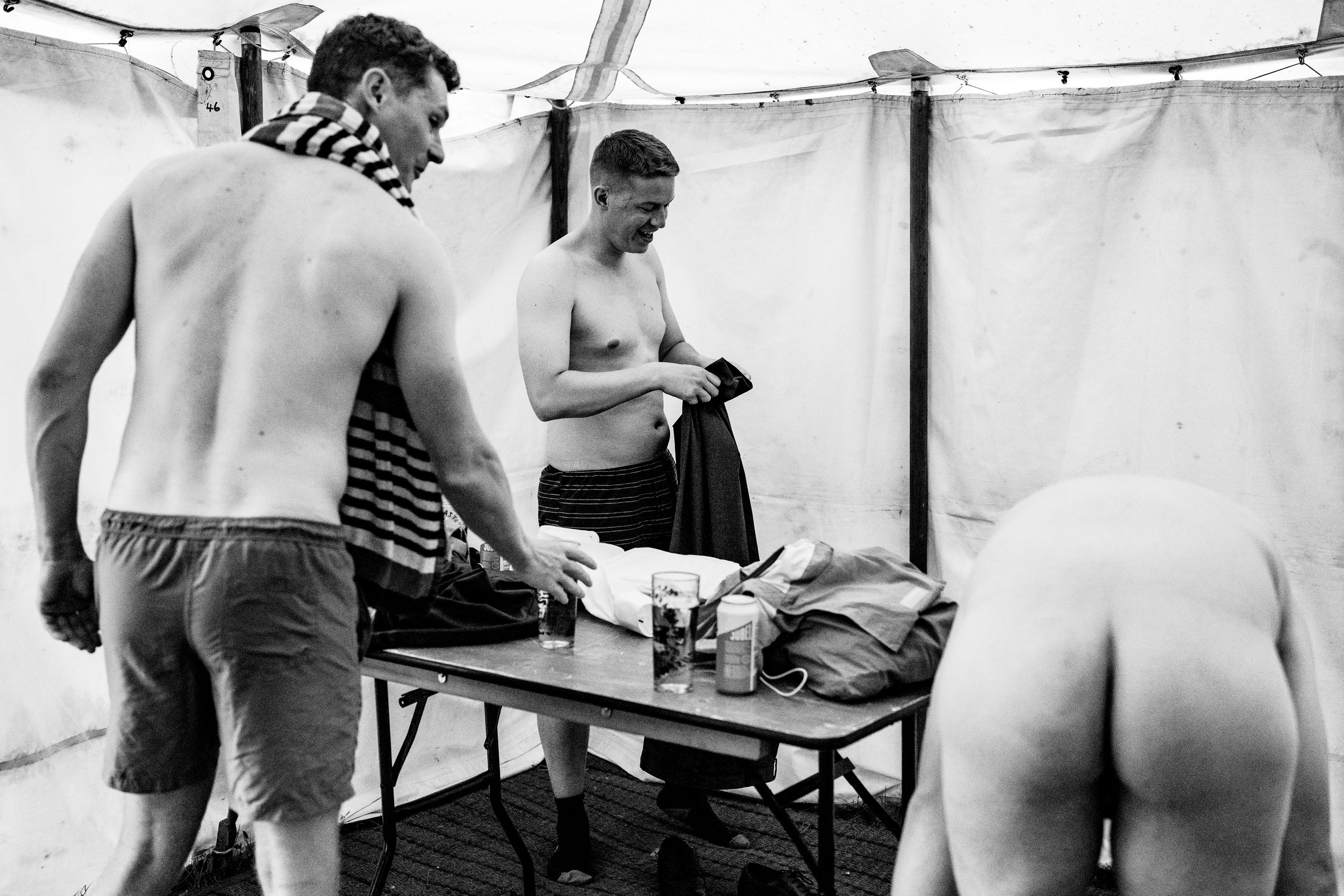 Black and white photo of shirtless men changing clothes in a tent at an outdoor event.