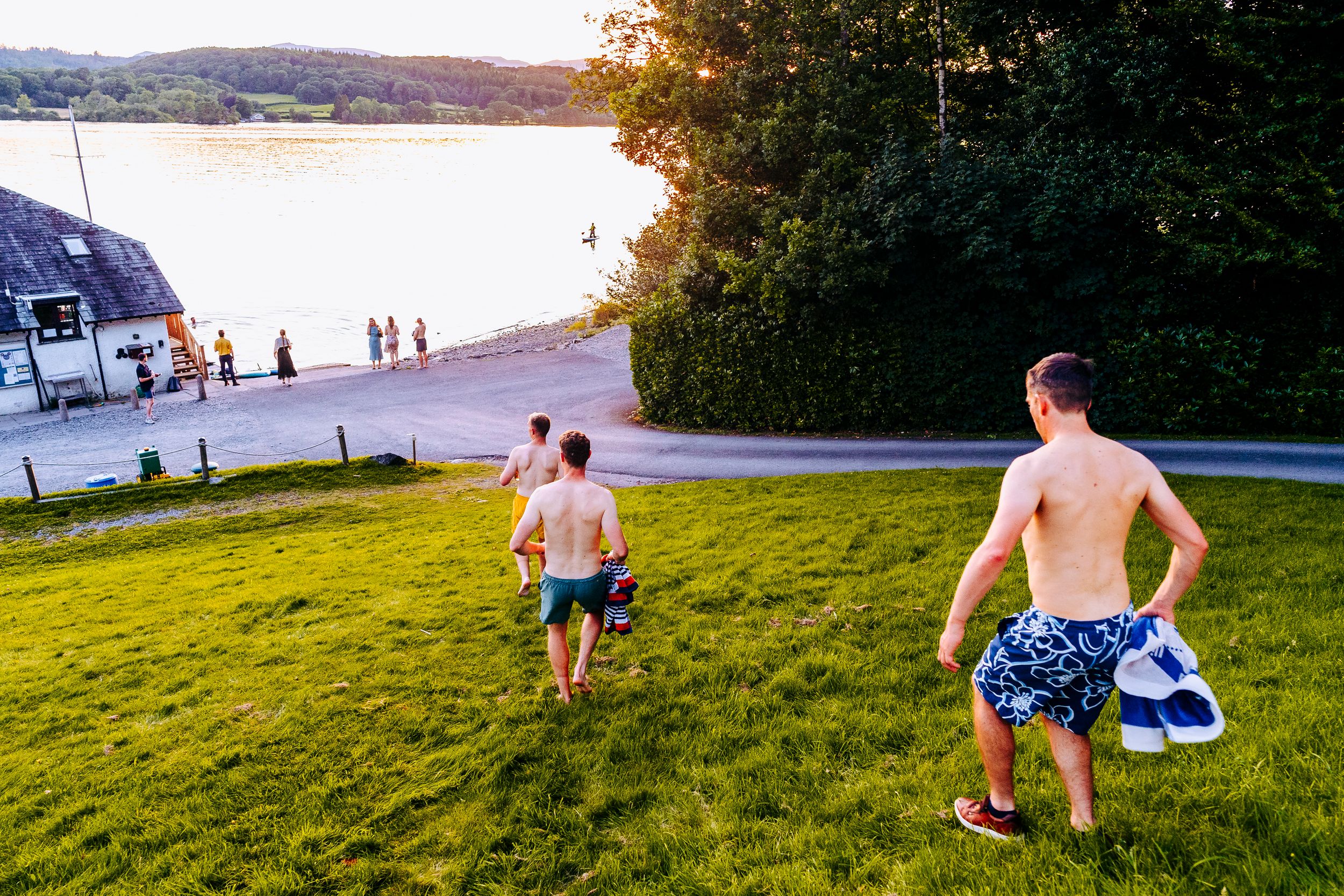 Two shirtless men walking toward a lakeside beach at sunset carrying towels.