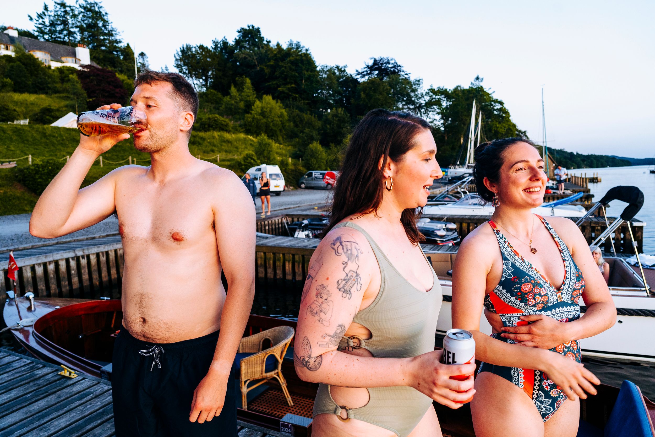 Three young adults enjoying drinks on a dock with boats and a lake in the background at dusk.