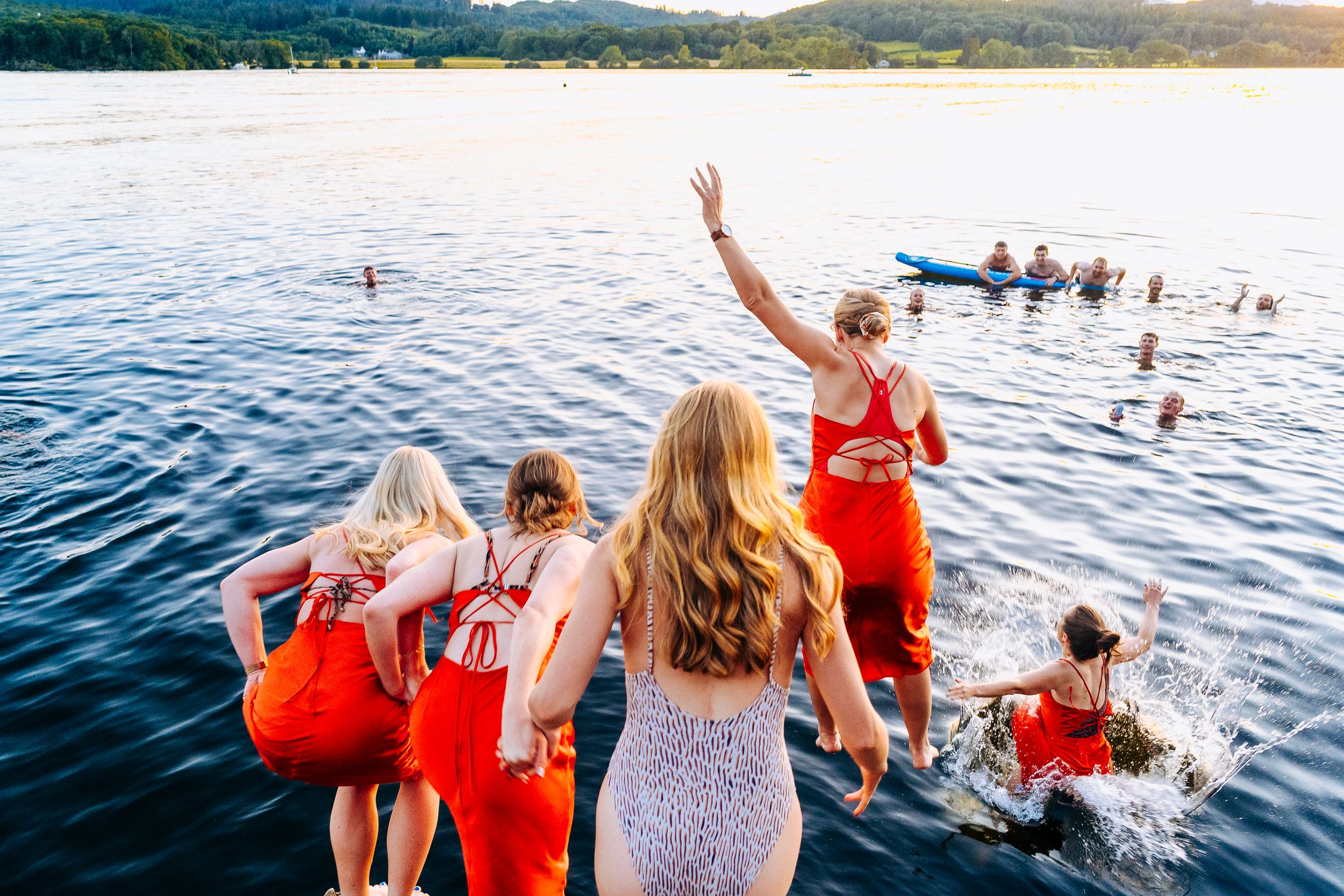 Women in red dresses jumping into a lake at sunset while others swim nearby.