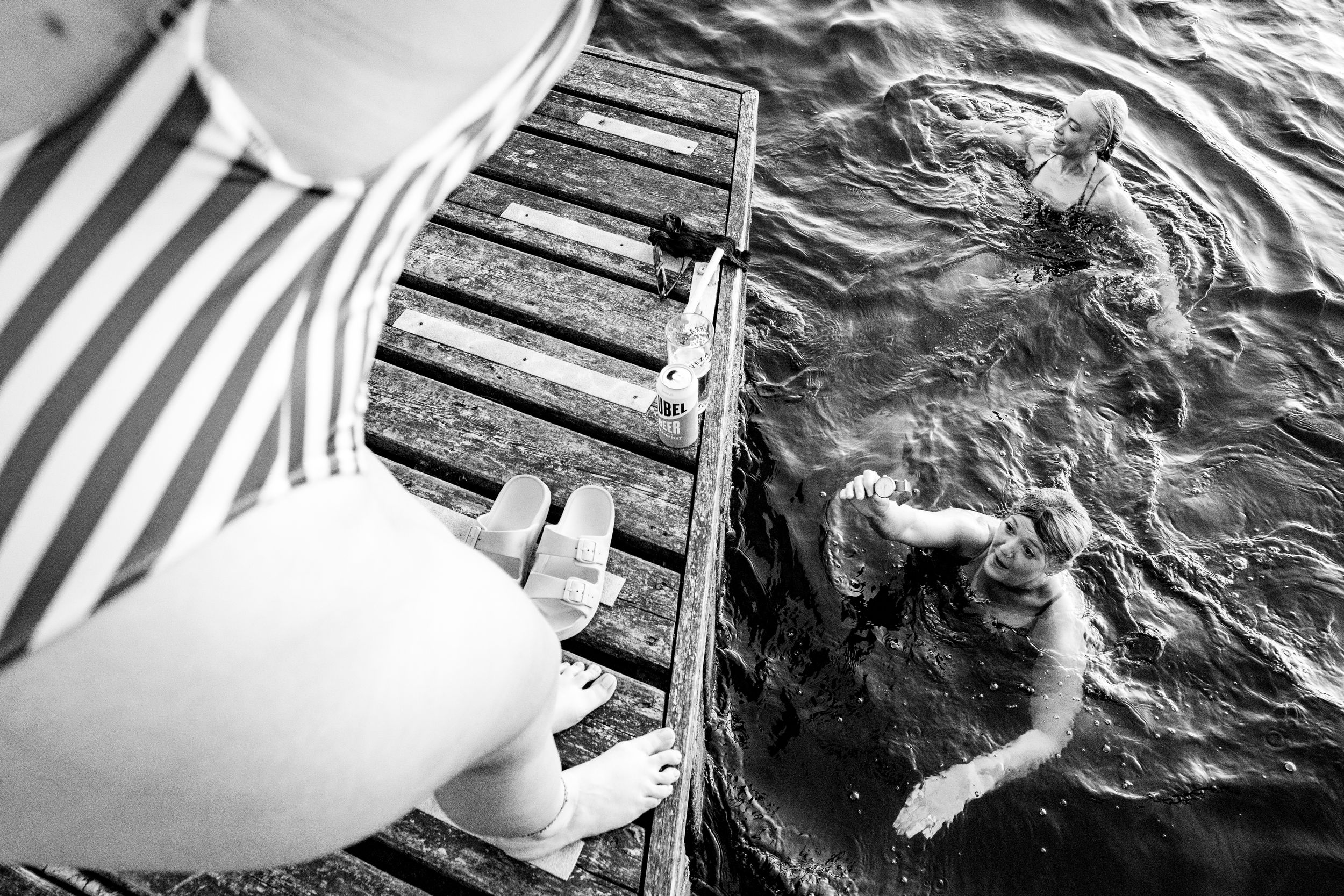 Black and white aerial view of a person sitting on a dock while someone swims below.