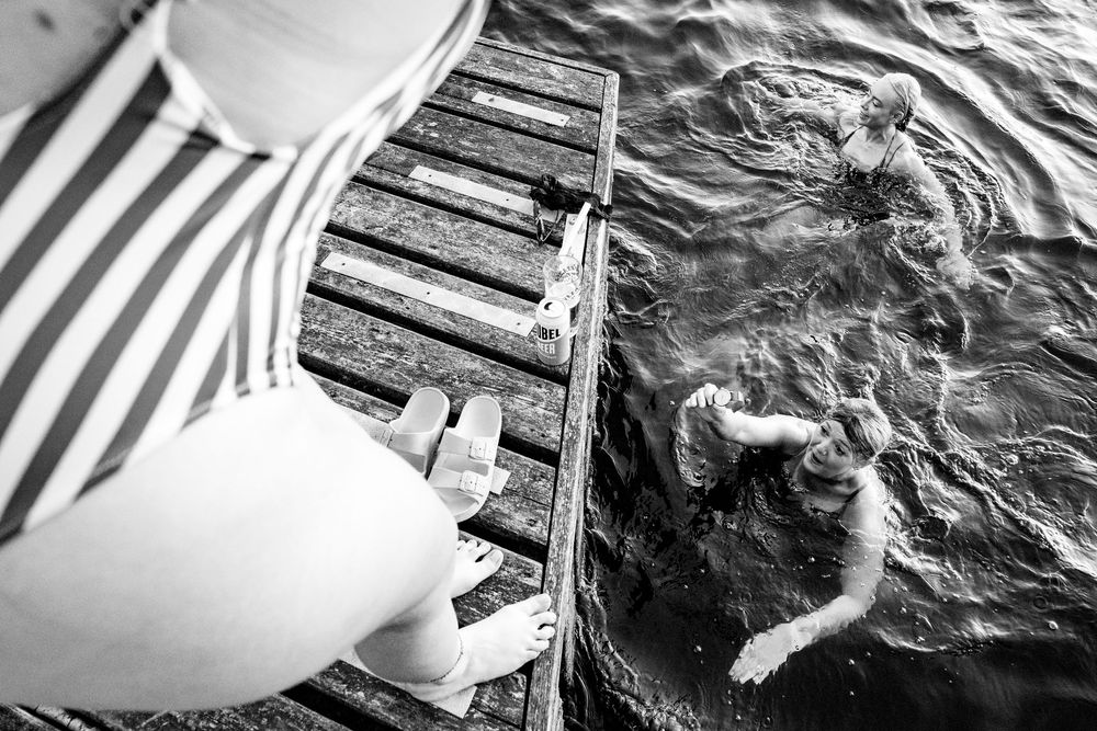 Black and white aerial view of a person sitting on a dock while someone swims below.