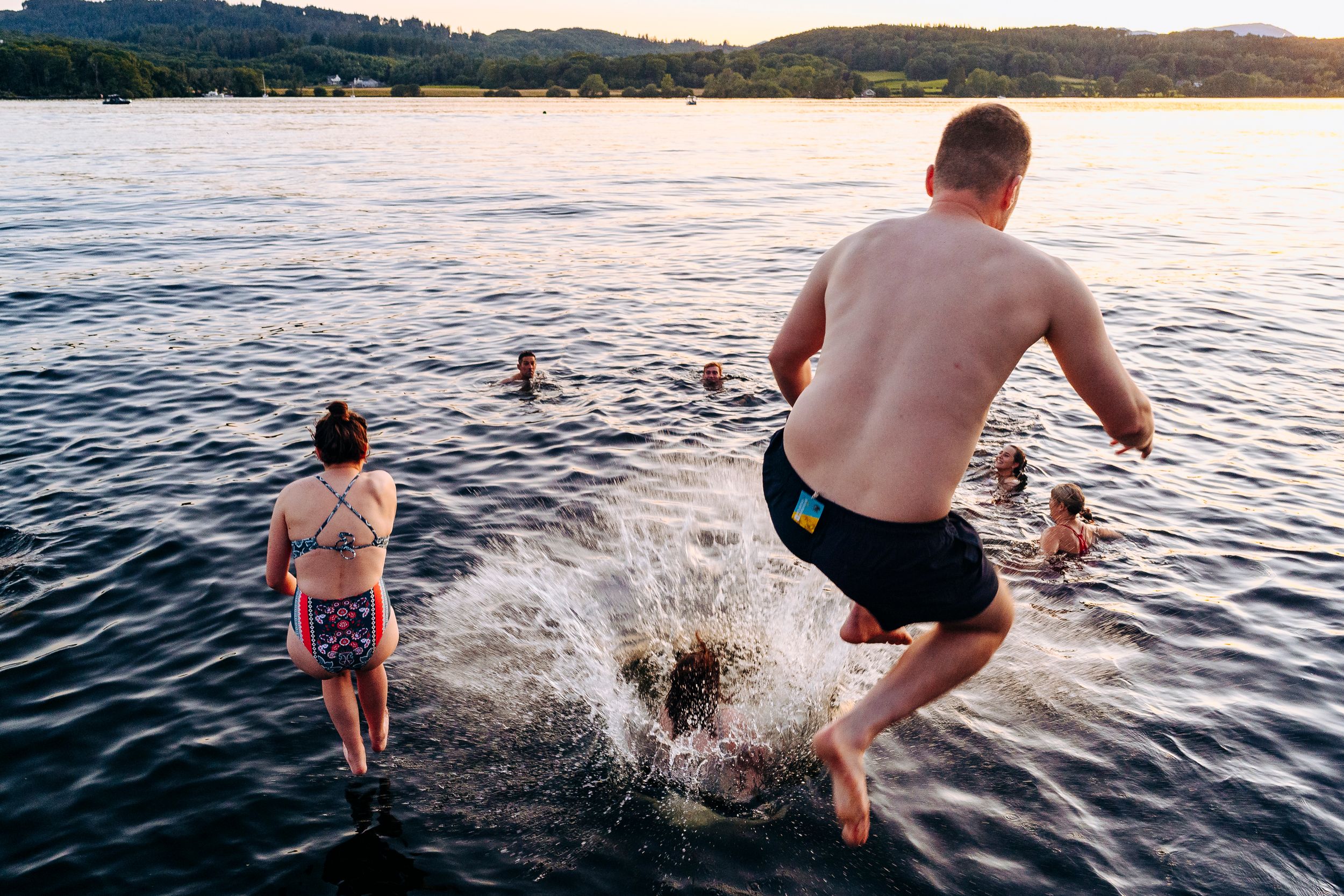 Shirtless man jumping into a lake at sunset while others swim in the background.