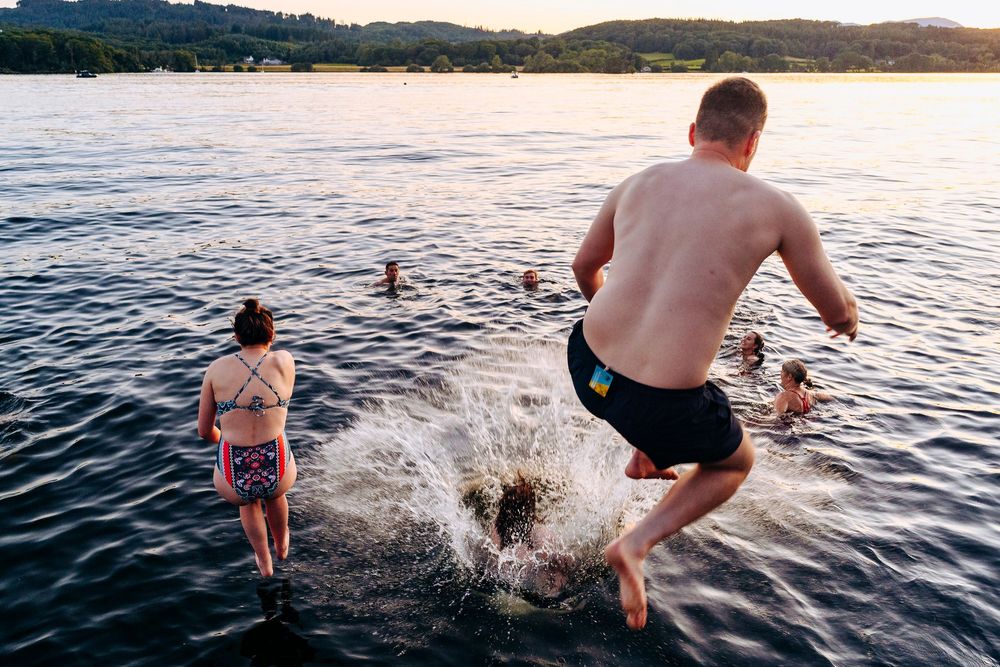 Shirtless man jumping into a lake at sunset while others swim in the background.