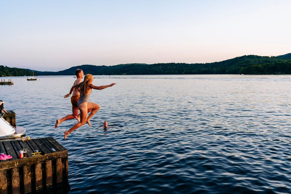 Bride and Groom leaping off a wooden dock into Lake Windermere at dusk in swimwear.