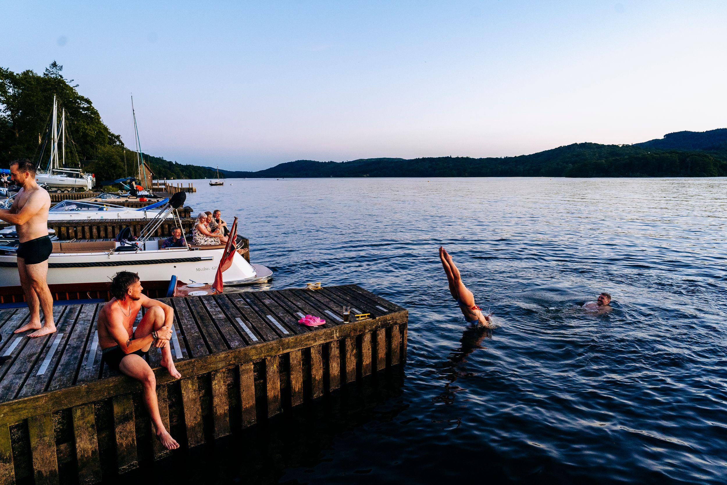 People swimming and diving off a wooden dock into a calm lake at dusk with sailboats moored nearby.