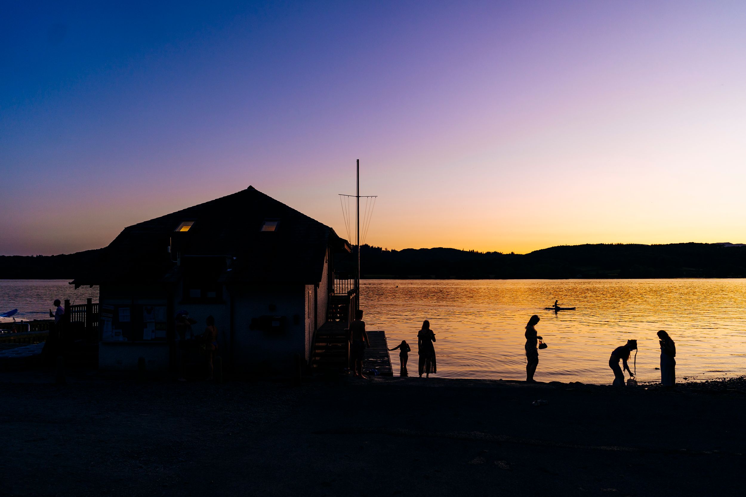 Silhouettes of people standing on a beach at sunset beside a boathouse with vibrant purple and orange sky in Windermere