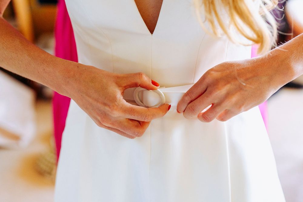 Bride having her white wedding dress belt fastened by a bridesmaid during wedding preparations.