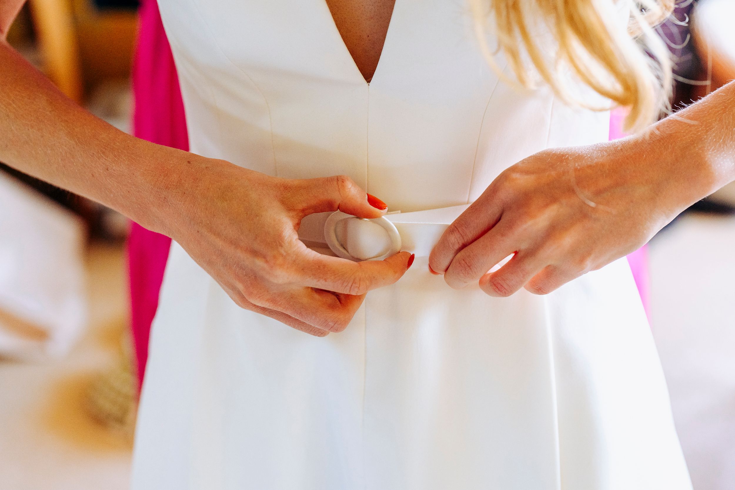 Bride having her white wedding dress belt fastened by a bridesmaid during wedding preparations.