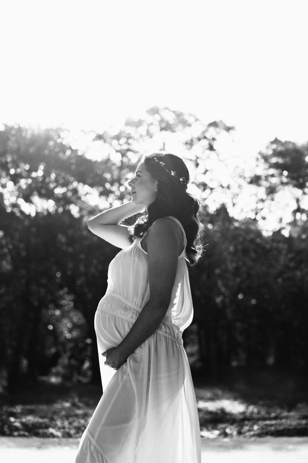 A silhouetted pregnant woman in a flowing white dress poses outdoors in black and white photography.