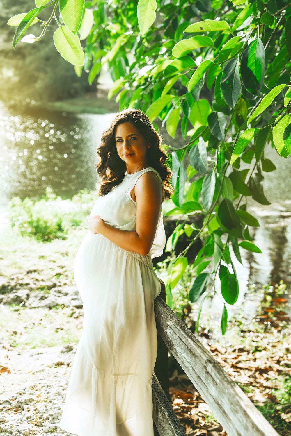 Expecting mother in flowing white dress poses by wooden fence near water and green foliage on sunny day.