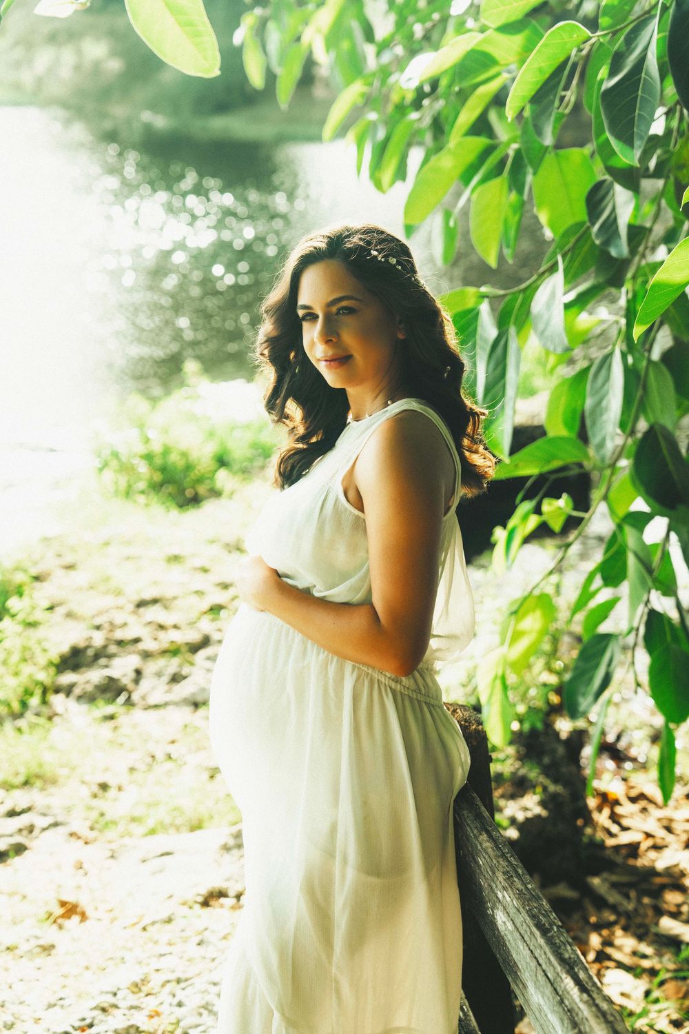 A person in a flowing white dress poses gracefully near lush green foliage in a sunlit outdoor setting.