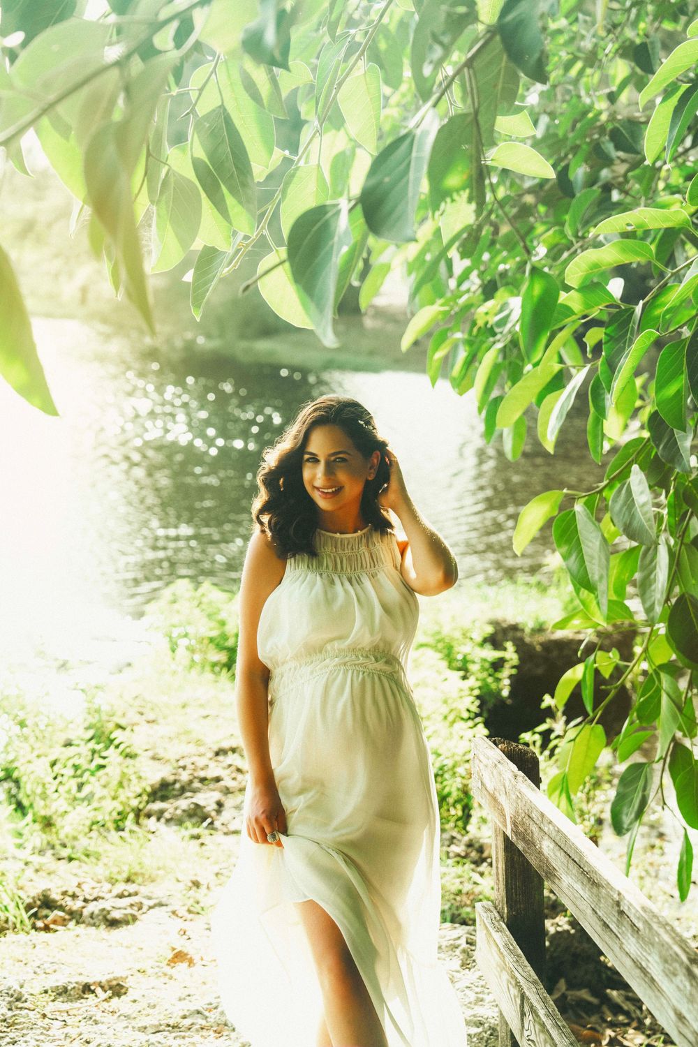 A person in a flowing white dress poses outdoors among green foliage and trees in bright sunlight.