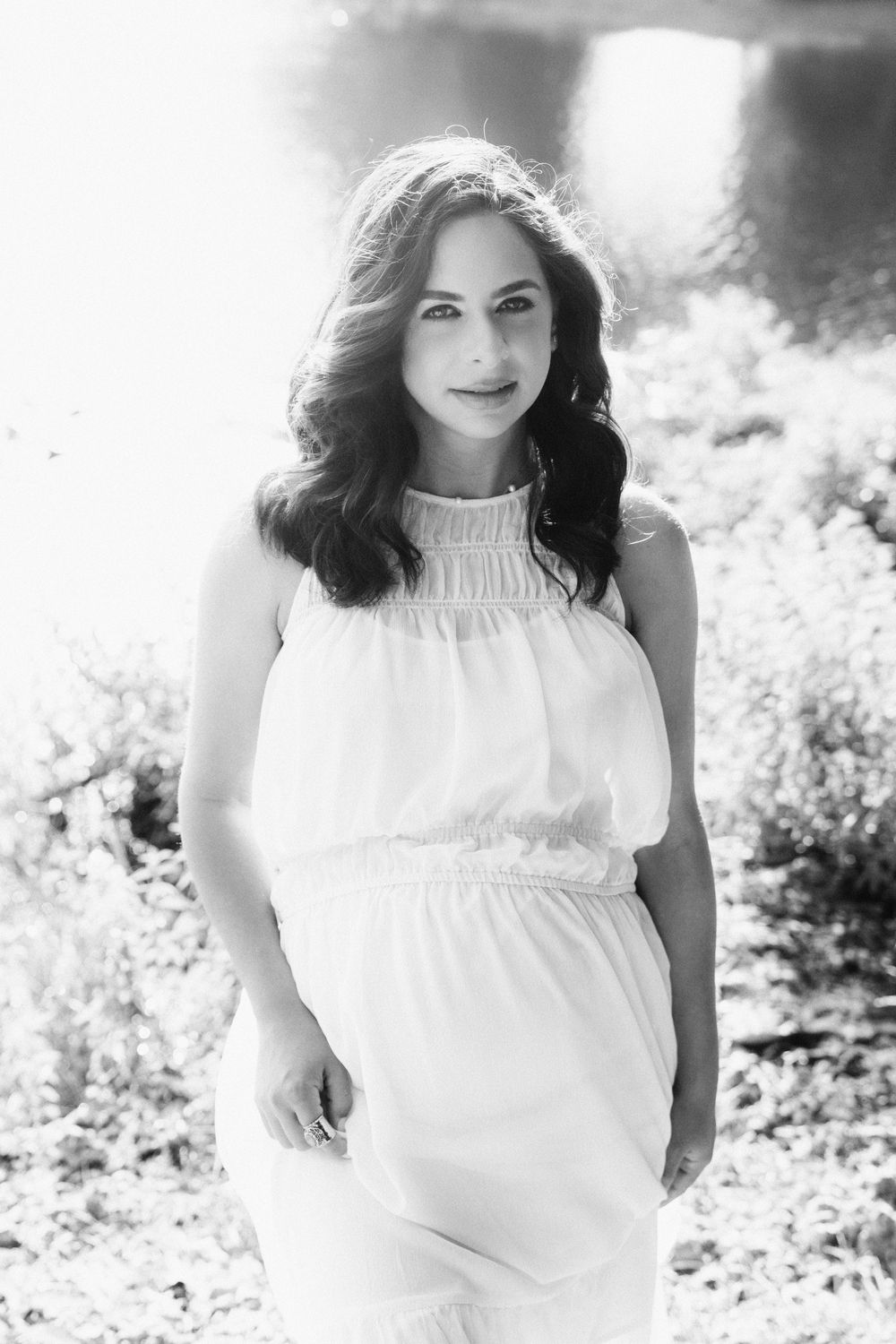 Black and white portrait of someone in a flowing white summer dress standing in dappled sunlight outdoors.