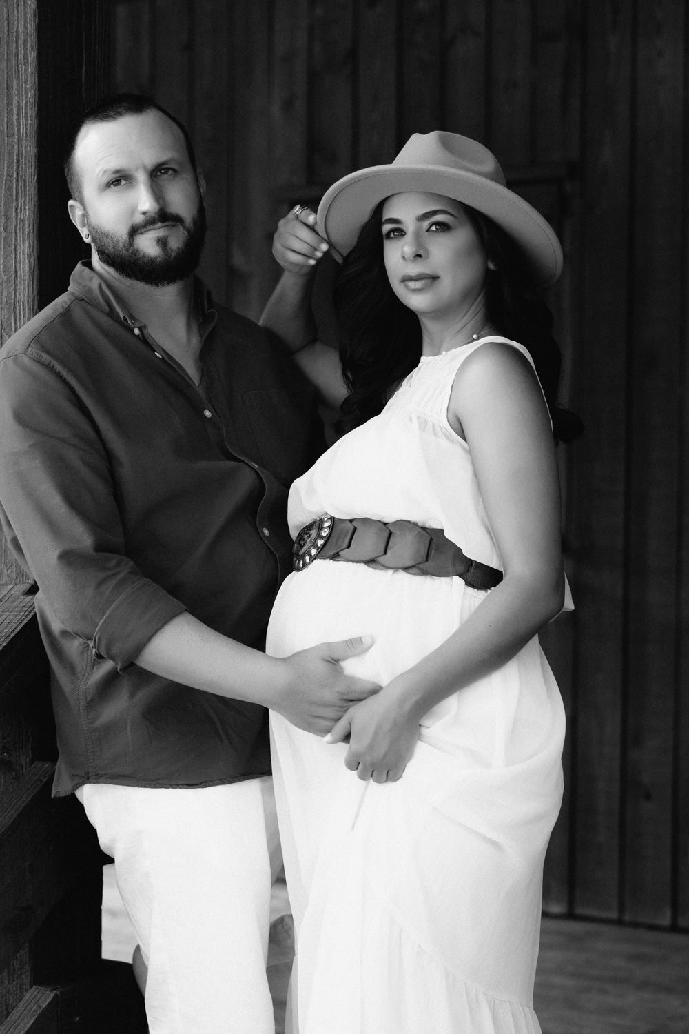 Black and white maternity photo of an expecting couple in white outfits posing against a wooden wall with rustic charm.