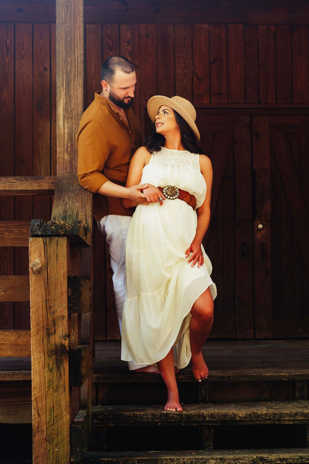 Expecting couple embraces on wooden dock stairs, woman in white dress and sun hat with partner in brown jacket.