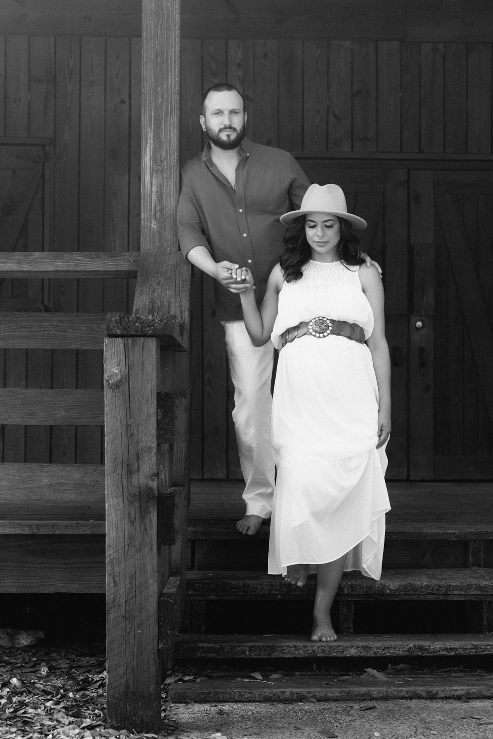 A couple stands on wooden stairs in black and white, with the woman wearing a flowing white dress and wide-brimmed hat.