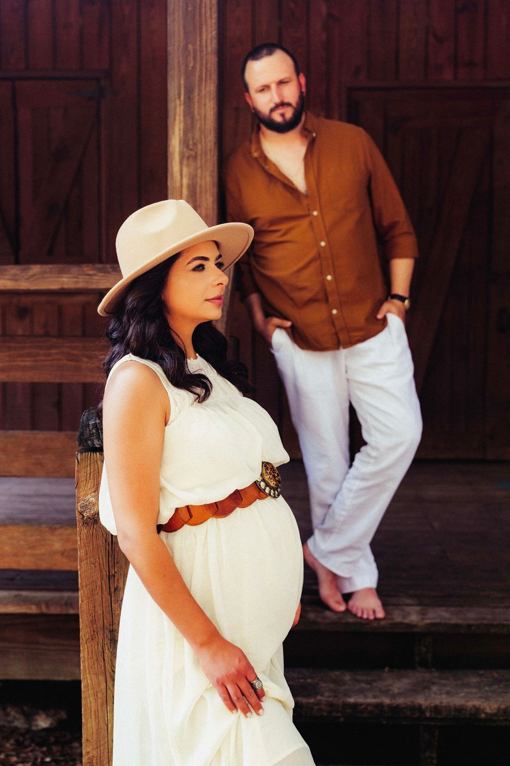 A couple pose for maternity photos by a rustic wooden barn, she in a white dress and hat, he in a brown button-down shirt.