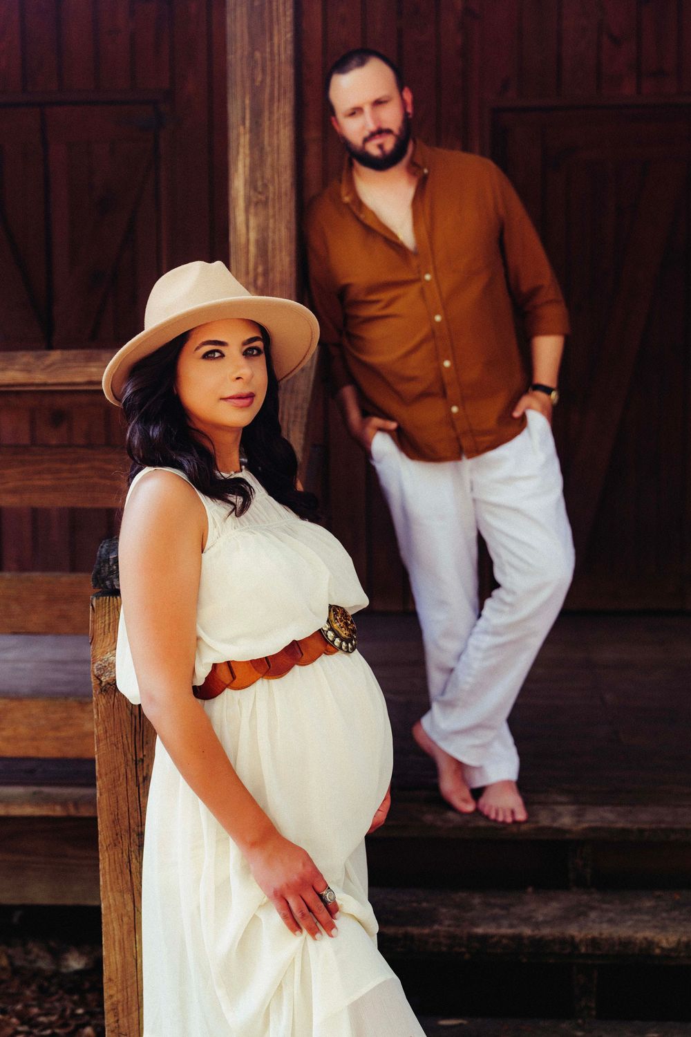 A couple poses on wooden steps for a maternity photo, with the pregnant woman in a white dress and sun hat.
