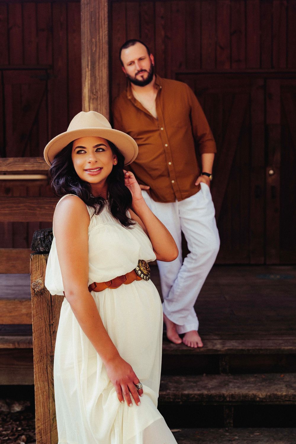 A couple poses against a rustic wooden backdrop, with a woman in white dress and sun hat and a man in brown shirt.