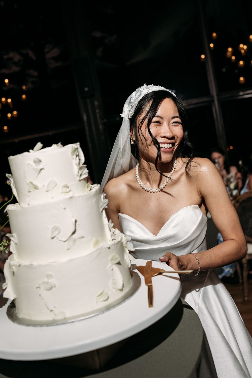 A bride in a white strapless gown and veil smiles joyfully while standing next to an elegant white tiered wedding cake.
