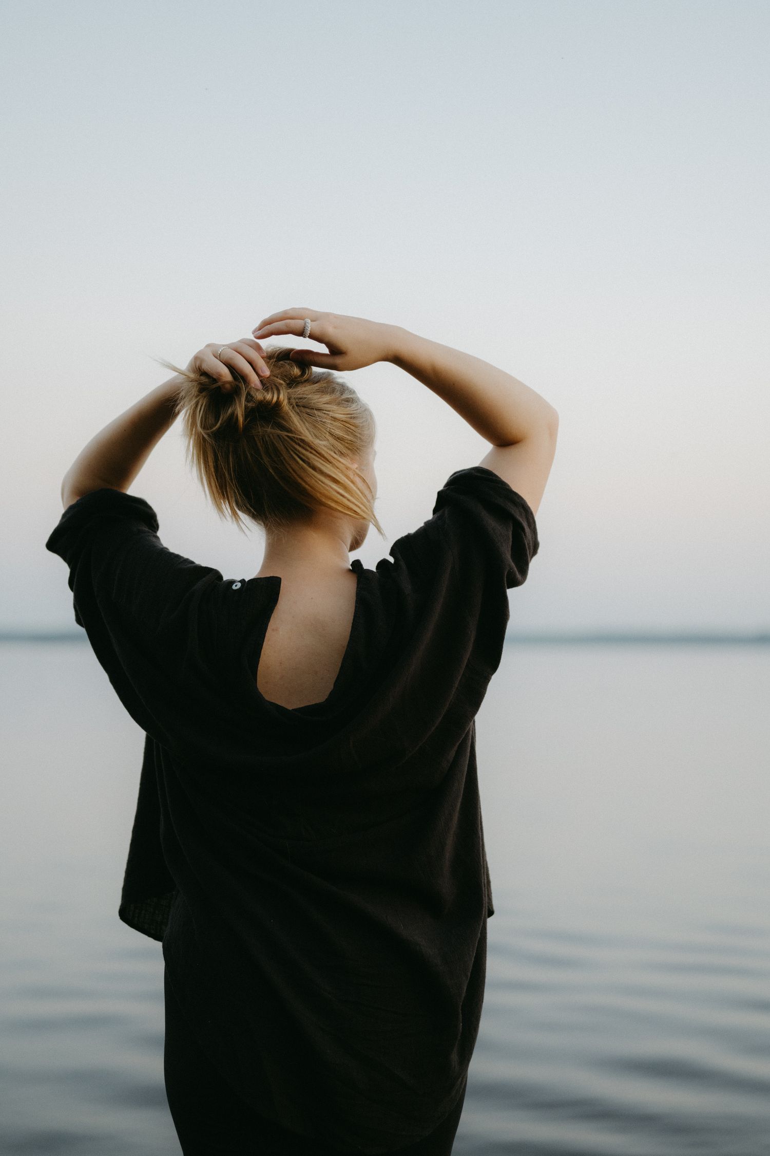Back view of person in black shirt adjusting hair while standing by calm water during sunset.