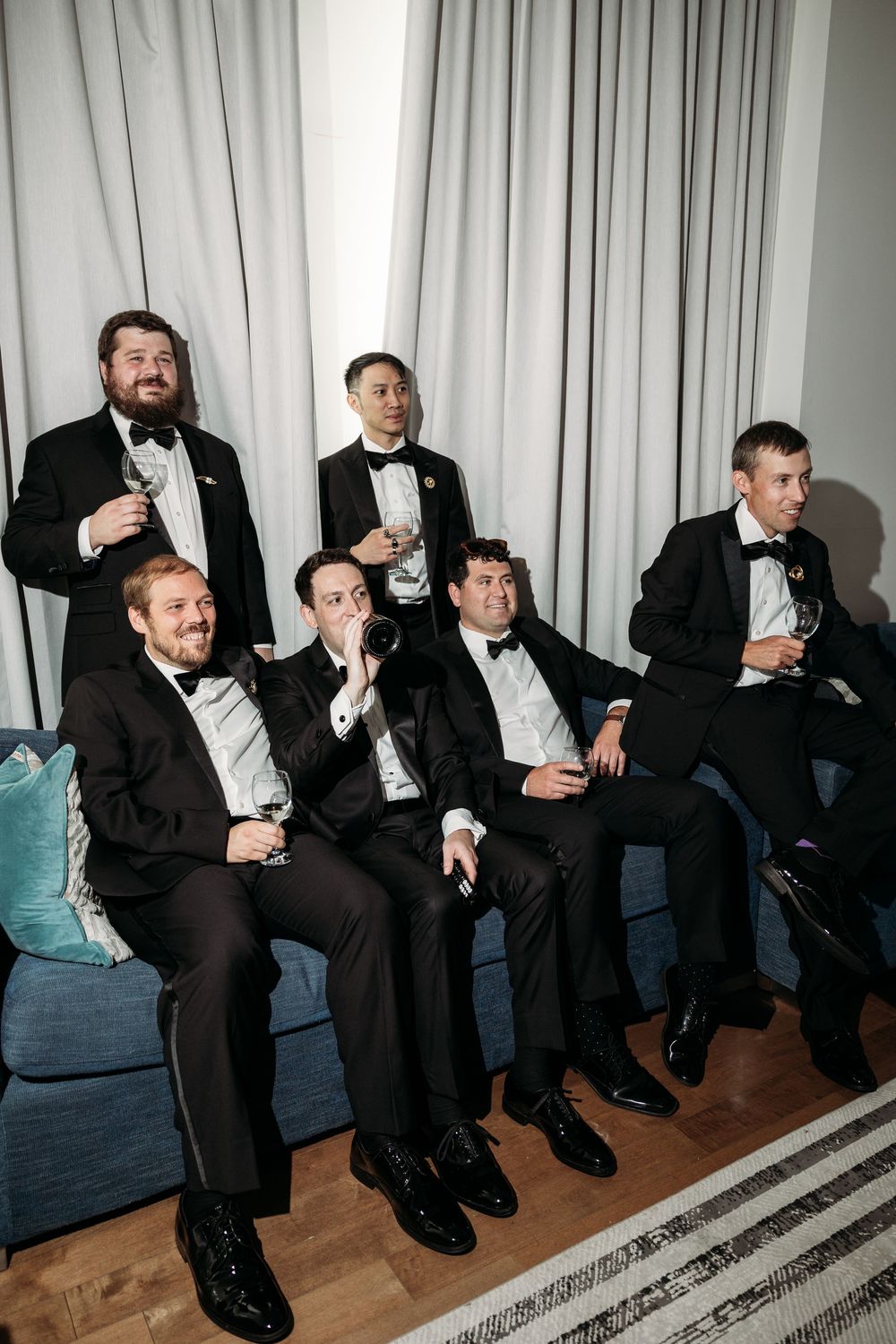 Groomsmen in black tuxedos and bowties relax on a blue couch at a formal wedding event.