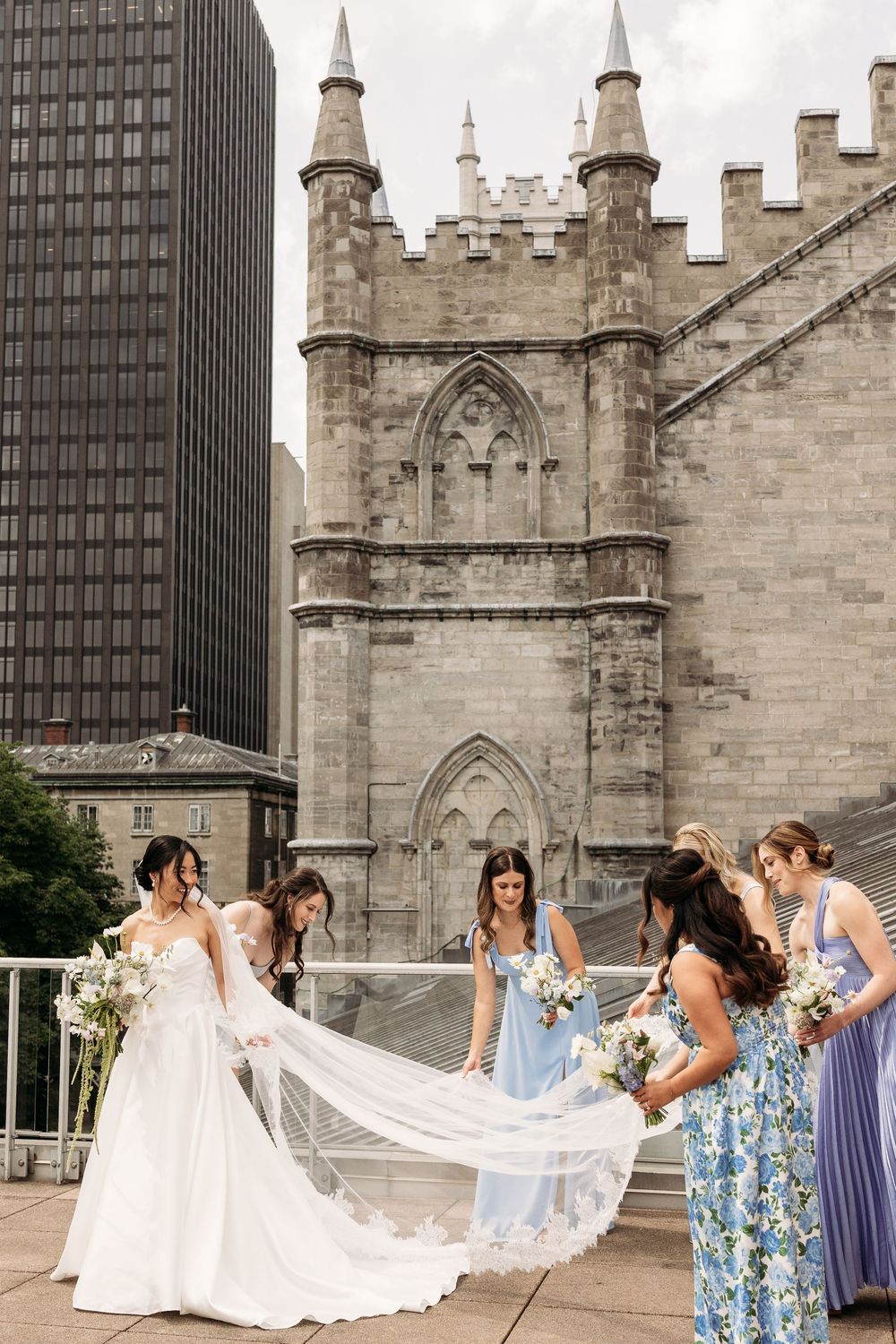 Bridal party in colorful dresses helps with white wedding gown veil in front of historic gothic church spires.