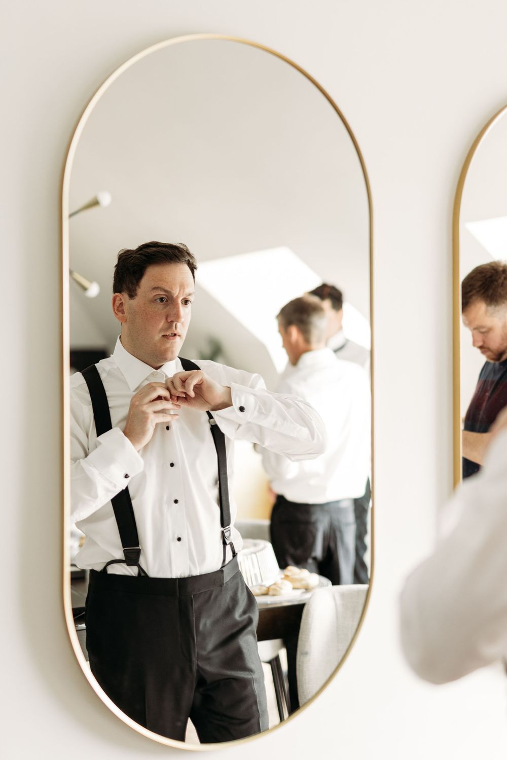 Elegant mirror reflection shows person adjusting formal attire with tuxedo shirt and suspenders while getting ready.