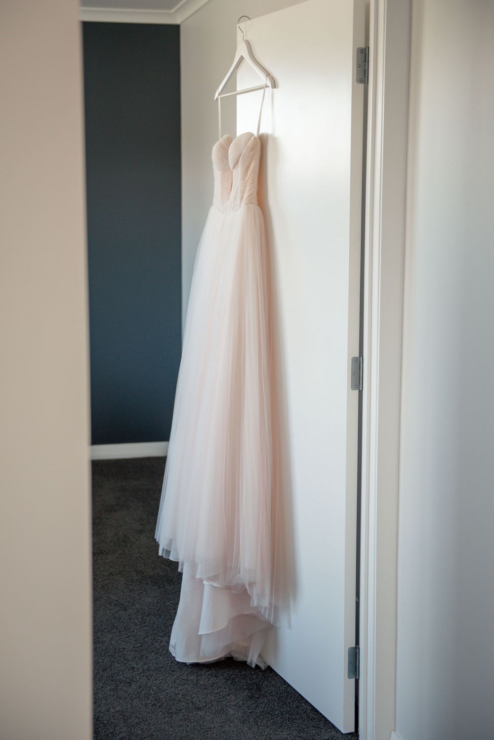 A blush tulle wedding gown hangs on display against a white wall with dark accent wall Cambridge New Zealand