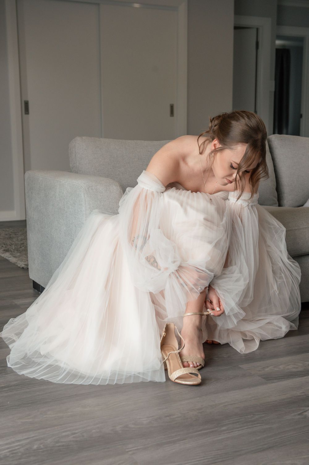 Bride in flowing white gown adjusts her strappy heels while seated on light gray couch Cambridge New Zealand