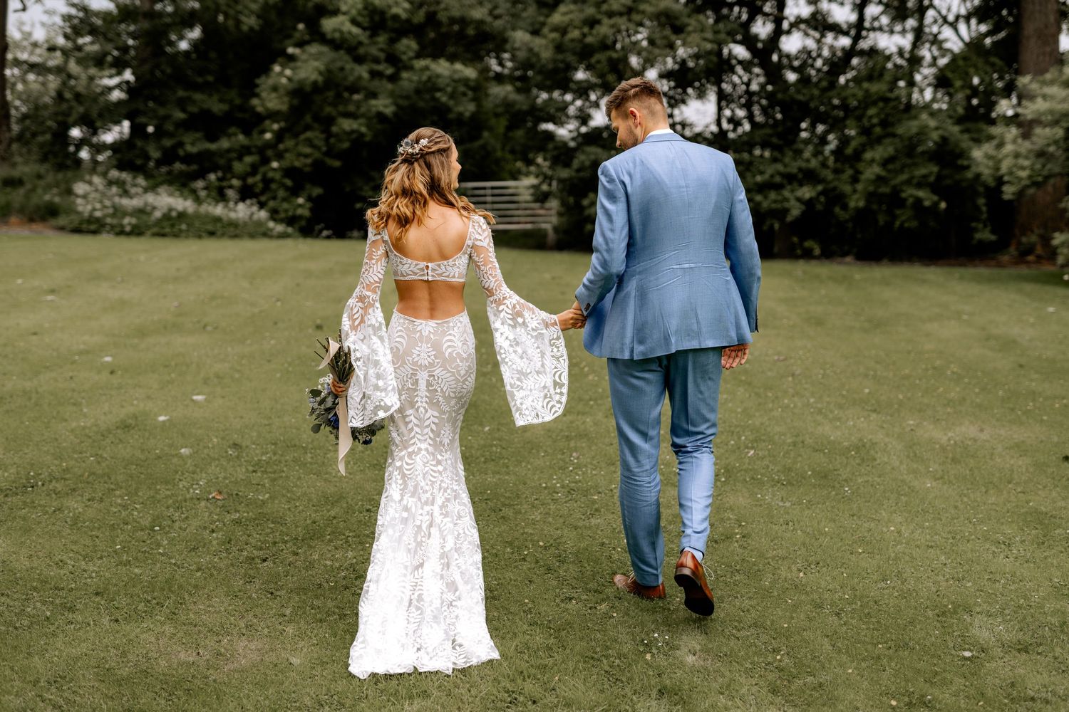 A couple walking away on green grass, with the bride wearing a fitted lace gown with bell sleeves.