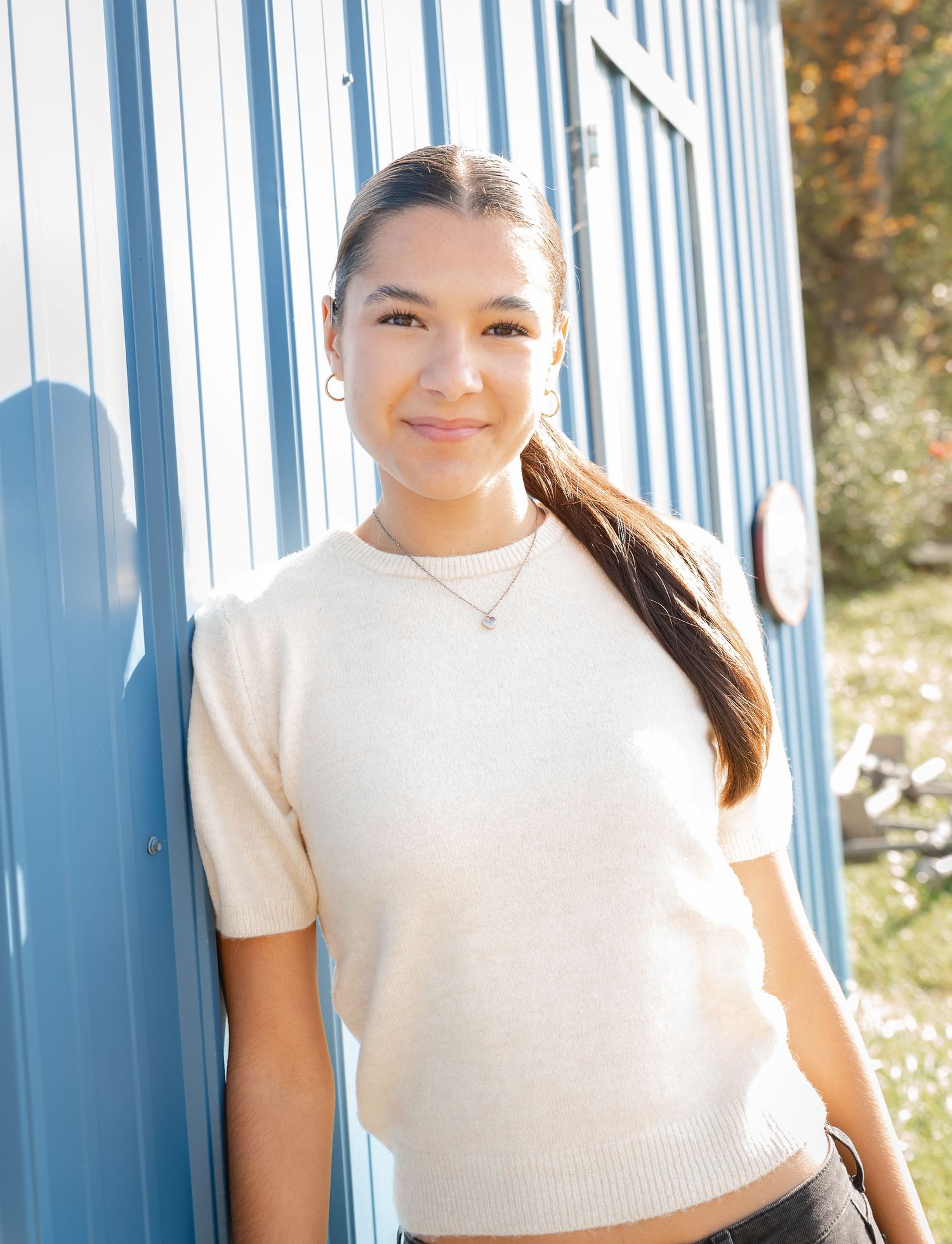 Someone in a cream sweater poses against a blue wall in natural lighting.