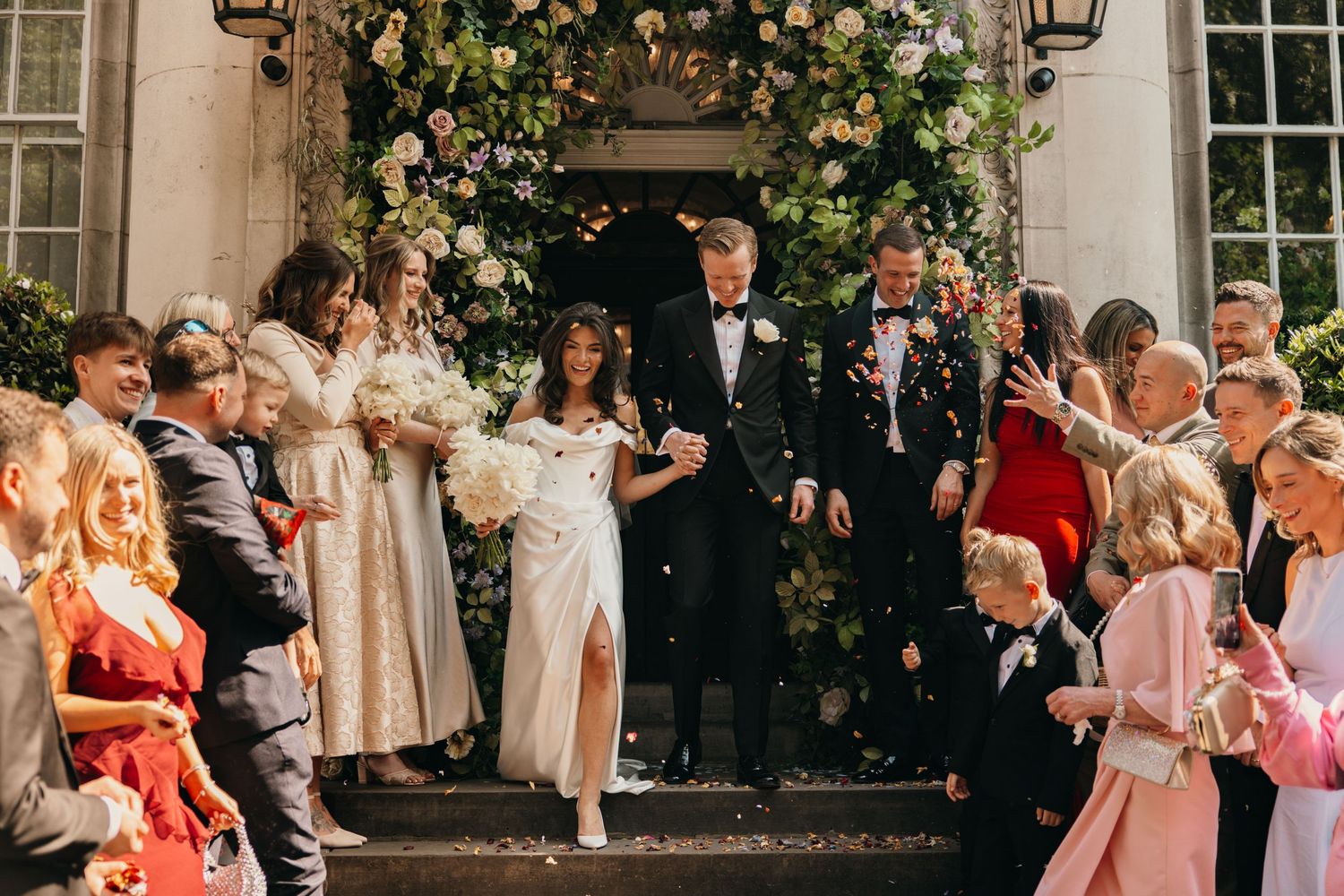 Wedding celebration on grand stone steps with ornate floral arch and cheering guests.