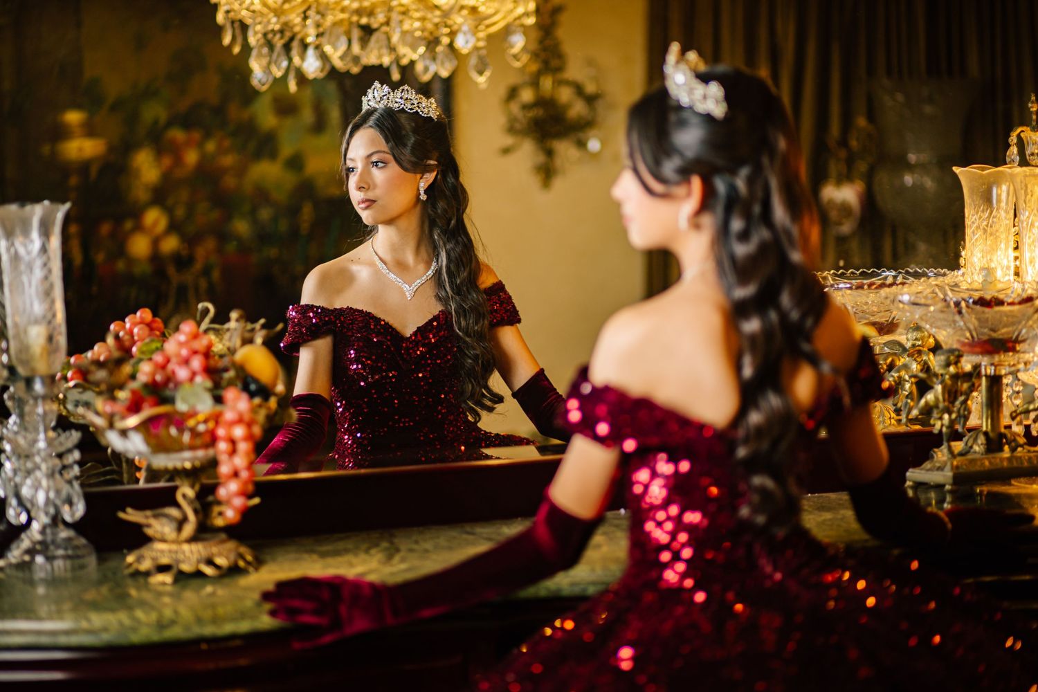 Elegant reflection in mirror shows sparkly burgundy ball gown with off-shoulder design amid luxurious decor and candlelight.