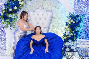 Two people pose in a luxurious setting with white throne chair and blue floral decorations.
