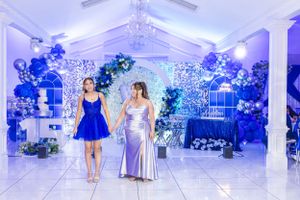 Two people in elegant dresses stand in a beautifully decorated venue with blue balloon arches and floral arrangements.