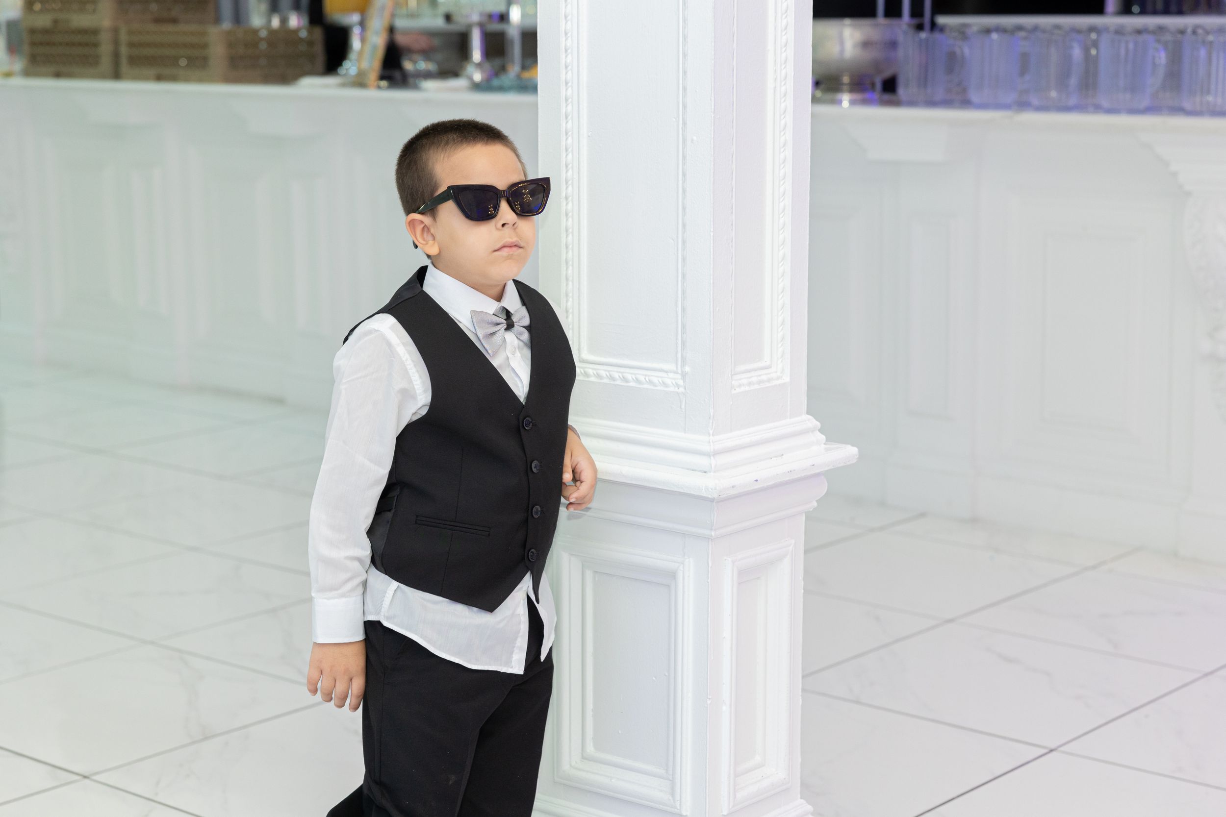 A young attendant in formal attire with sunglasses poses beside a white column.