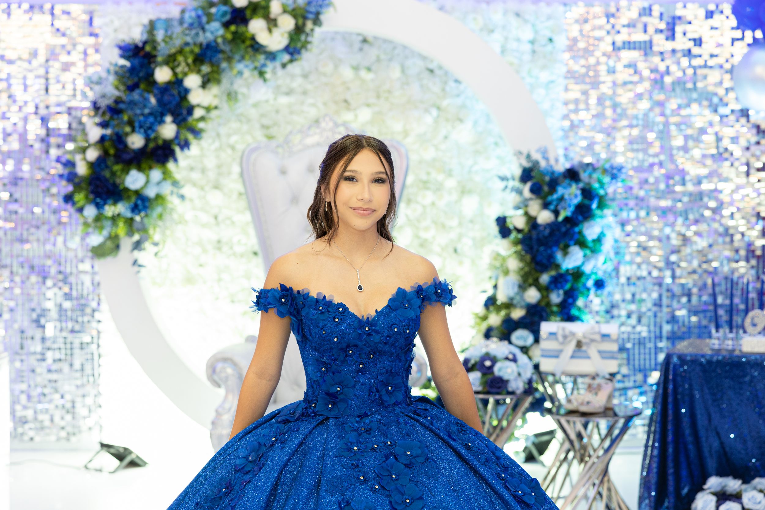 A royal blue ball gown sparkles in an elegant white and blue decorated venue with floral accents.