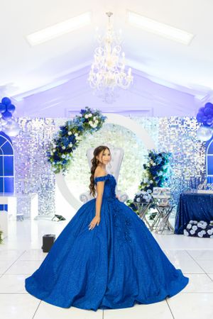 An elegant blue ball gown shimmers in a beautifully decorated white venue with crystal chandeliers.