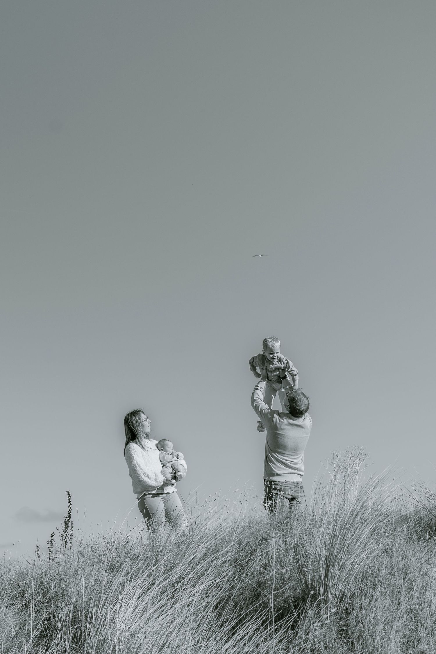 A black and white photograph capturing a tender family moment in a field of tall grass.