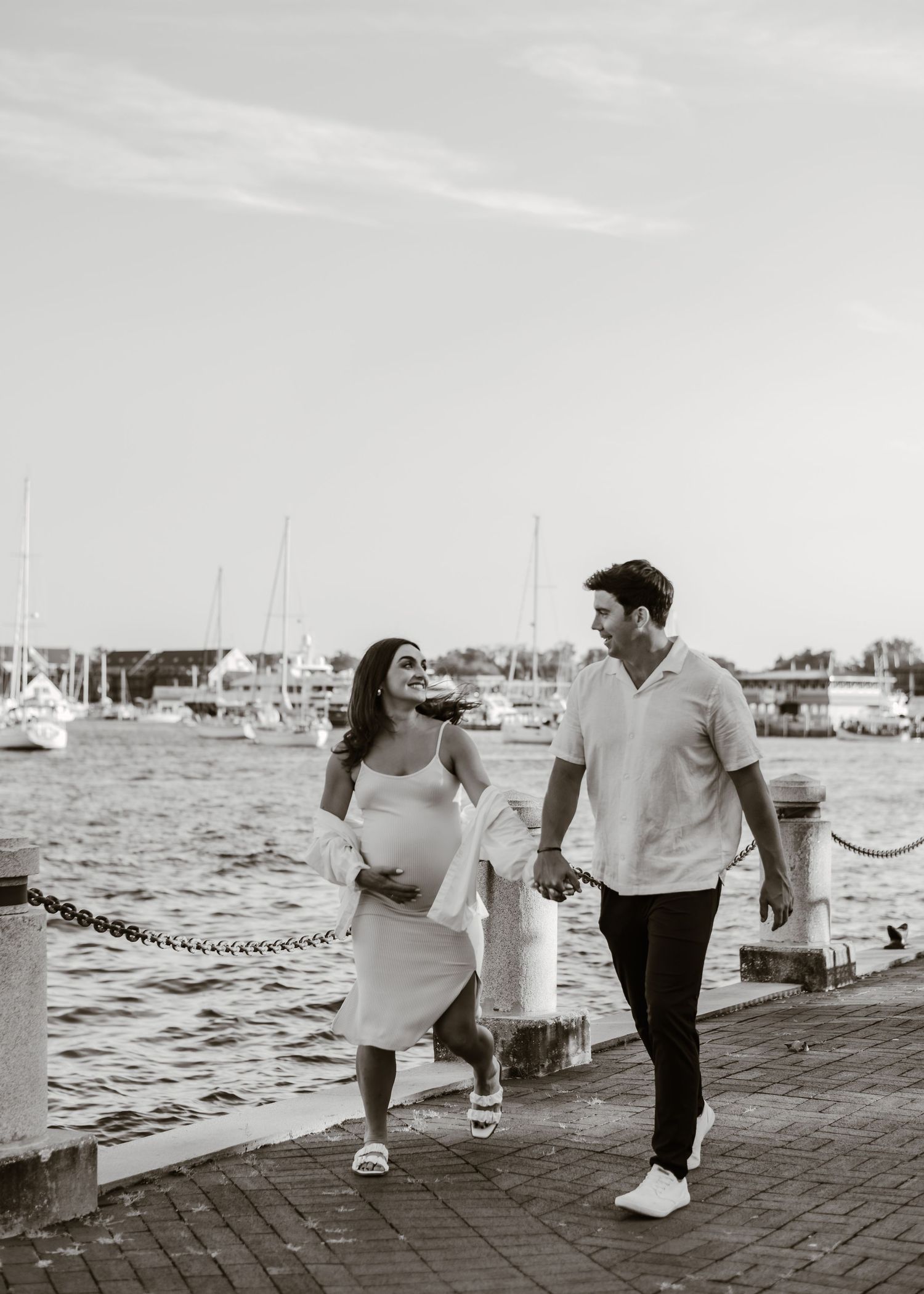 Couple walking hand in hand along a waterfront boardwalk with boats in the background.